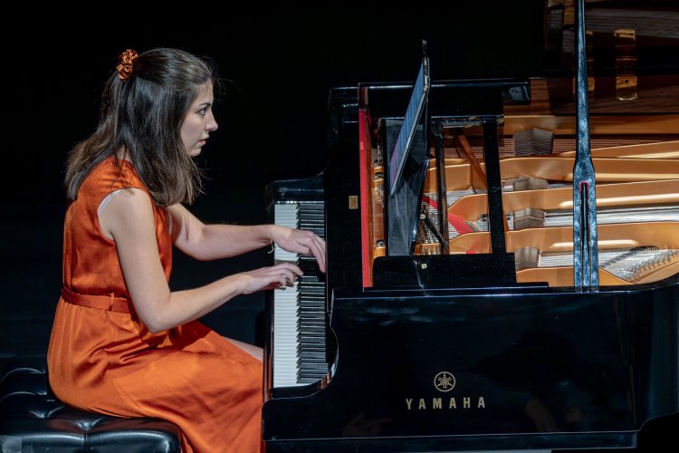 Female student wearing orange dress playing piano on stage.