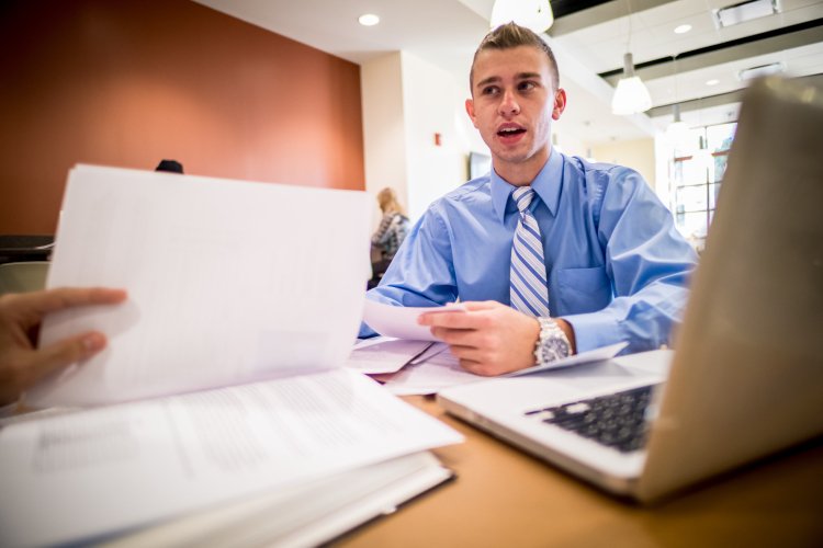 A student in a dress shirt and tie meets across a table covered with papers and a laptop.