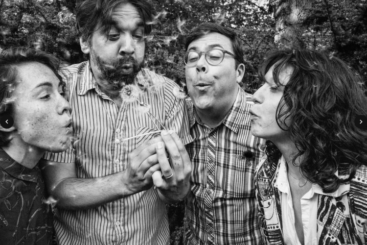 Members of The Hands Free blowing air at a dandelion