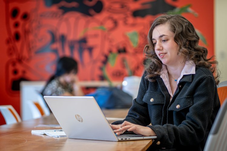 A student works on a laptop at a long table in a colorful study space with a bright mural on the wall behind her.