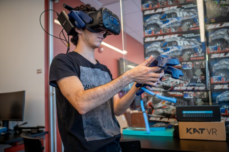 Student wearing a virtual reality headset uses handheld controllers in a lab filled with VR and gaming equipment.