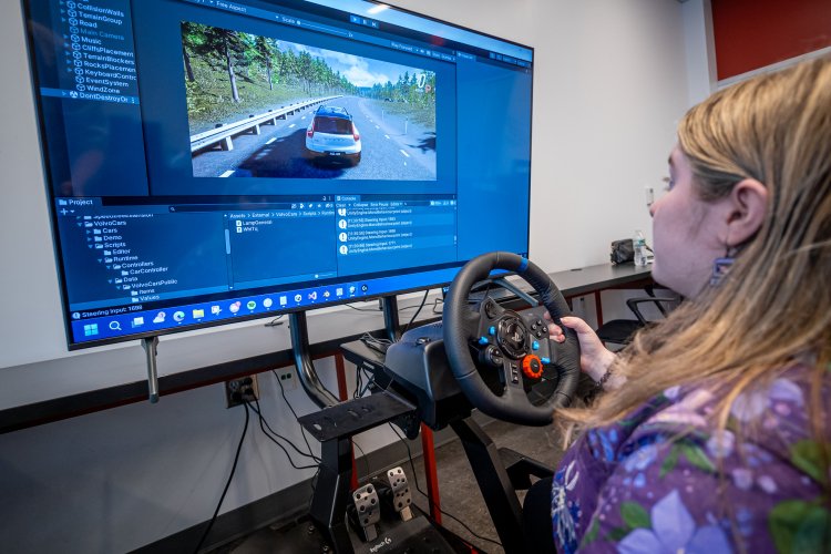 Student uses a steering wheel controller to test a driving simulation game displayed on a large monitor showing the game development software interface.