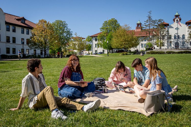 Students sit together on a blanket on a large grassy lawn at Montclair State University, talking and studying with notebooks and a laptop open, while academic buildings with red-tiled roofs and a clear blue sky frame the background.