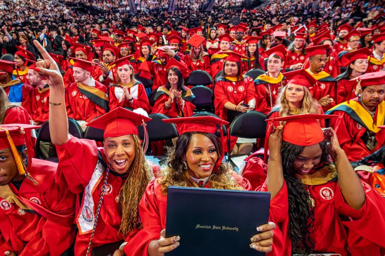 A large group of Montclair State University Graduate School students in bright red caps and gowns smile, cheer and hold up diplomas while seated together on the arena floor during the 2026 Spring Commencement ceremony, with rows of fellow graduates and guests filling the stands behind them.