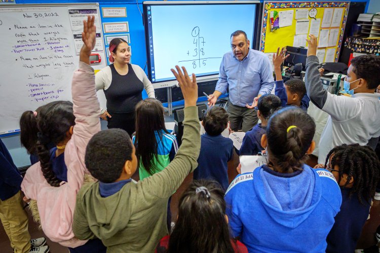 Two teachers at front of classroom surrounded by children with hands raised
