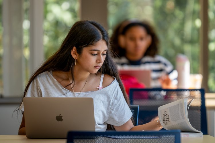 A student turns the pages to a book while working on her laptop in the library. A student wearing headphones sits behind her.