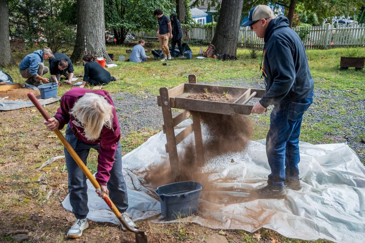 At a backyard archaeological dig, a woman digs a hole in the ground while a man uses a soil screener and others work in the background.