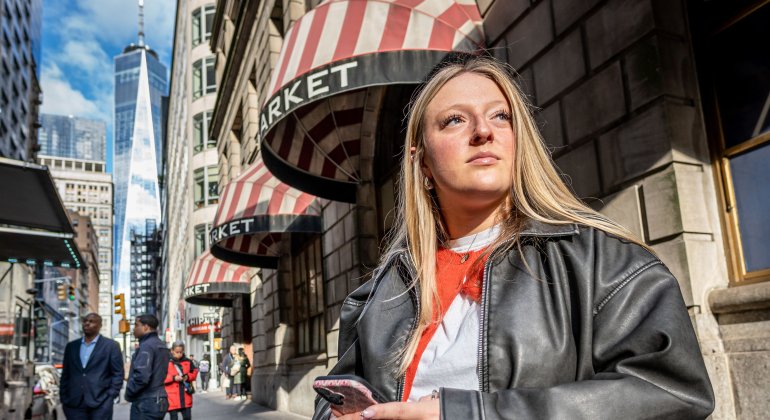 Emma Haskell stands outside a New York City subway stop, holding her phone, with a cityscape visible in the background.