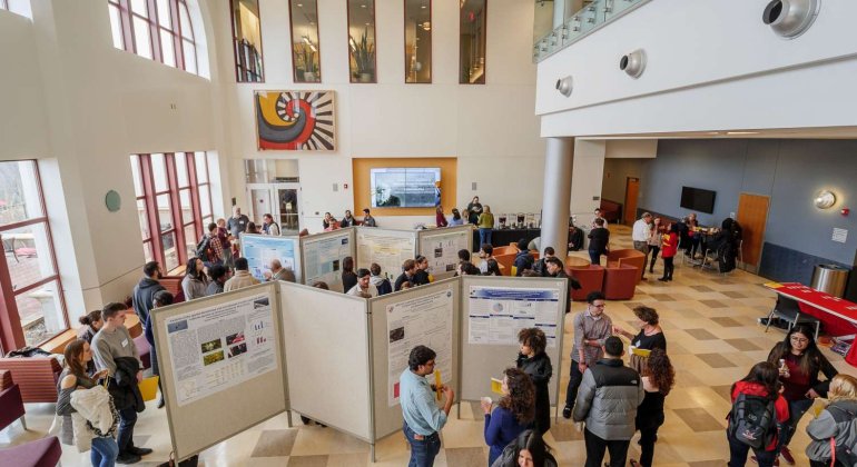 Wide view of the College of Environmental and Life Sciences atrium with student presentations arrayed on movable walls and a crowd of students and faculty moving around the space.