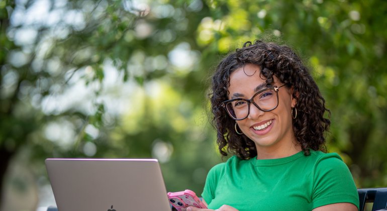 A student sits outside on a bench in a park setting while working on a laptop.
