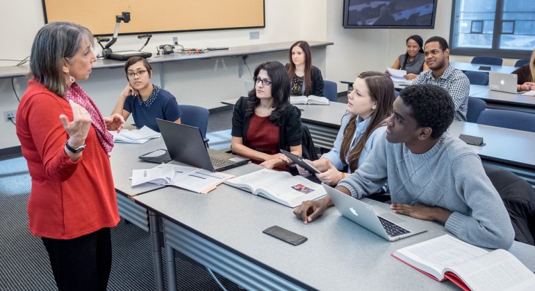 Professor leads a discussion with graduate students seated around a classroom table, laptops and open books in front of them as they engage in a group conversation.