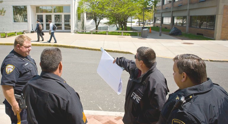 A montclair fire saftey officer holds blueprints and points toward a building exit while consulting with University police officers and a Passaic County Sherrif's Office officer