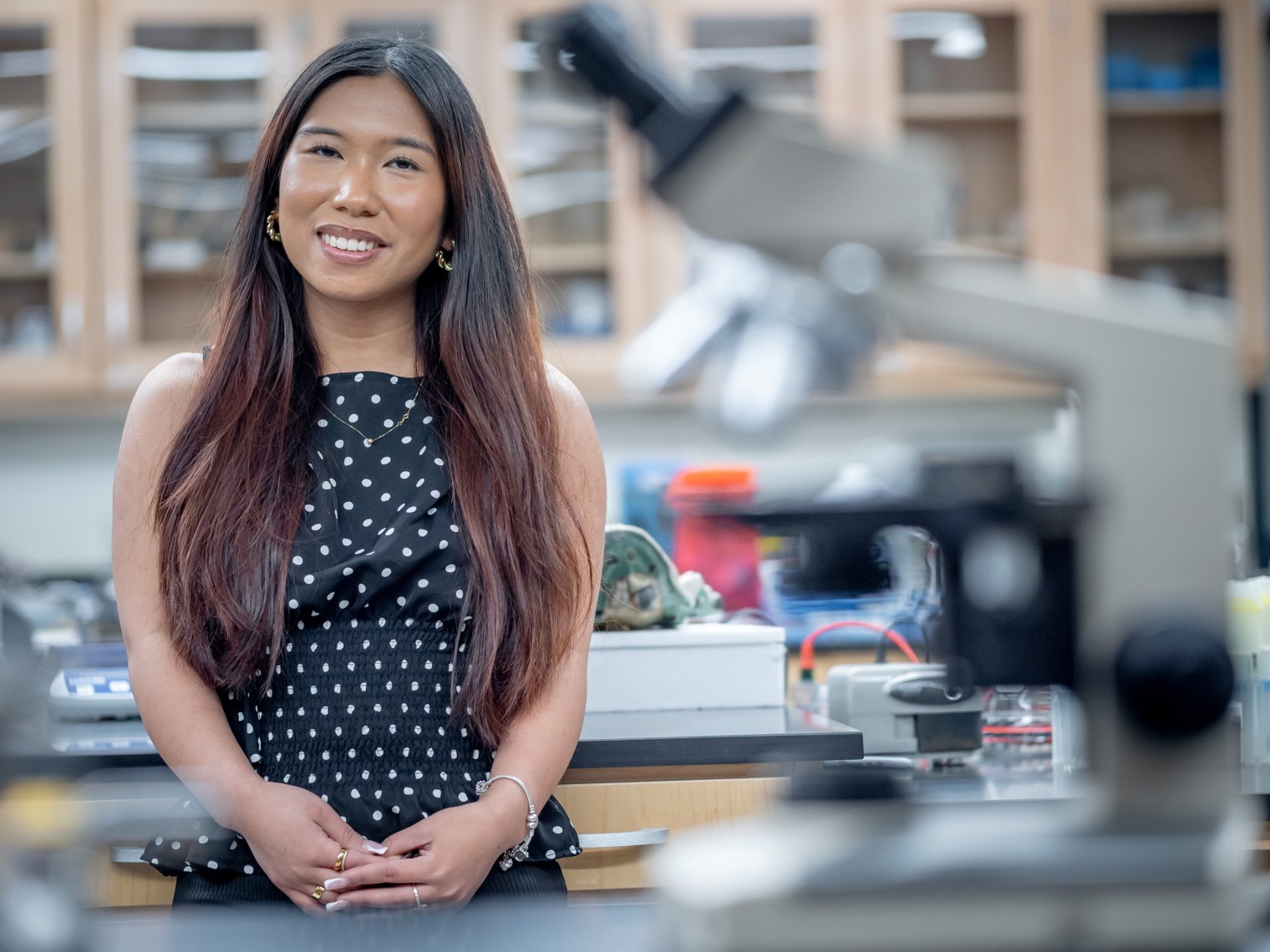 Woman is smiling, standing, next to a microscope. 