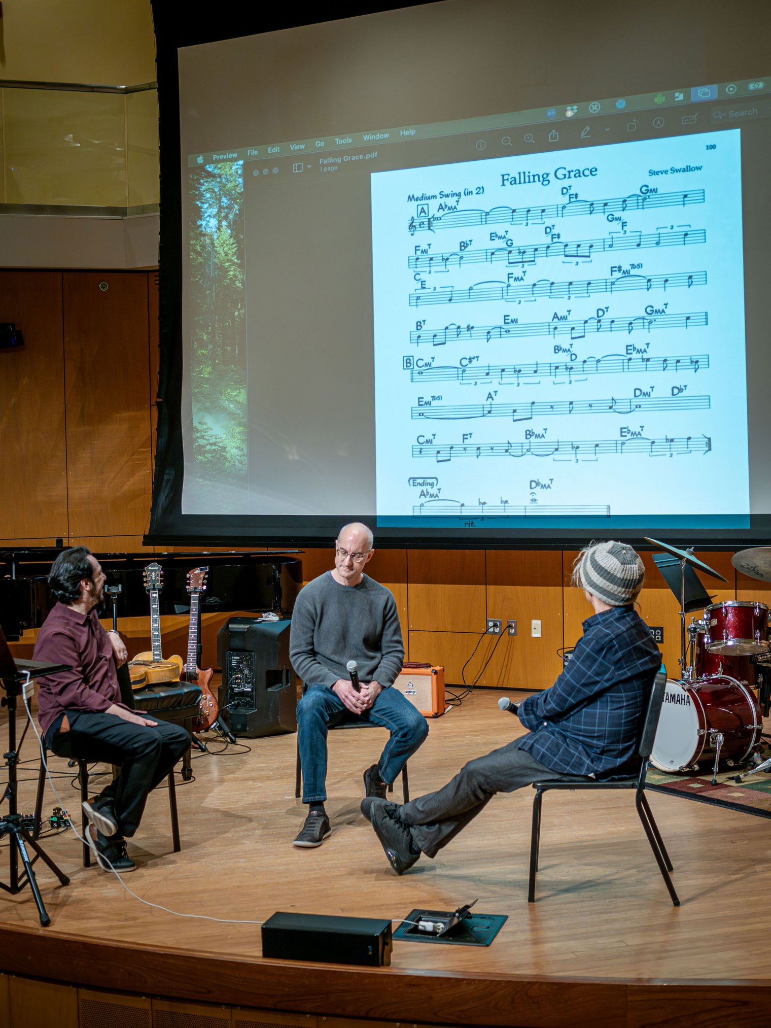 Three men onstage discuss a sheet of music projected overhead on a screen.