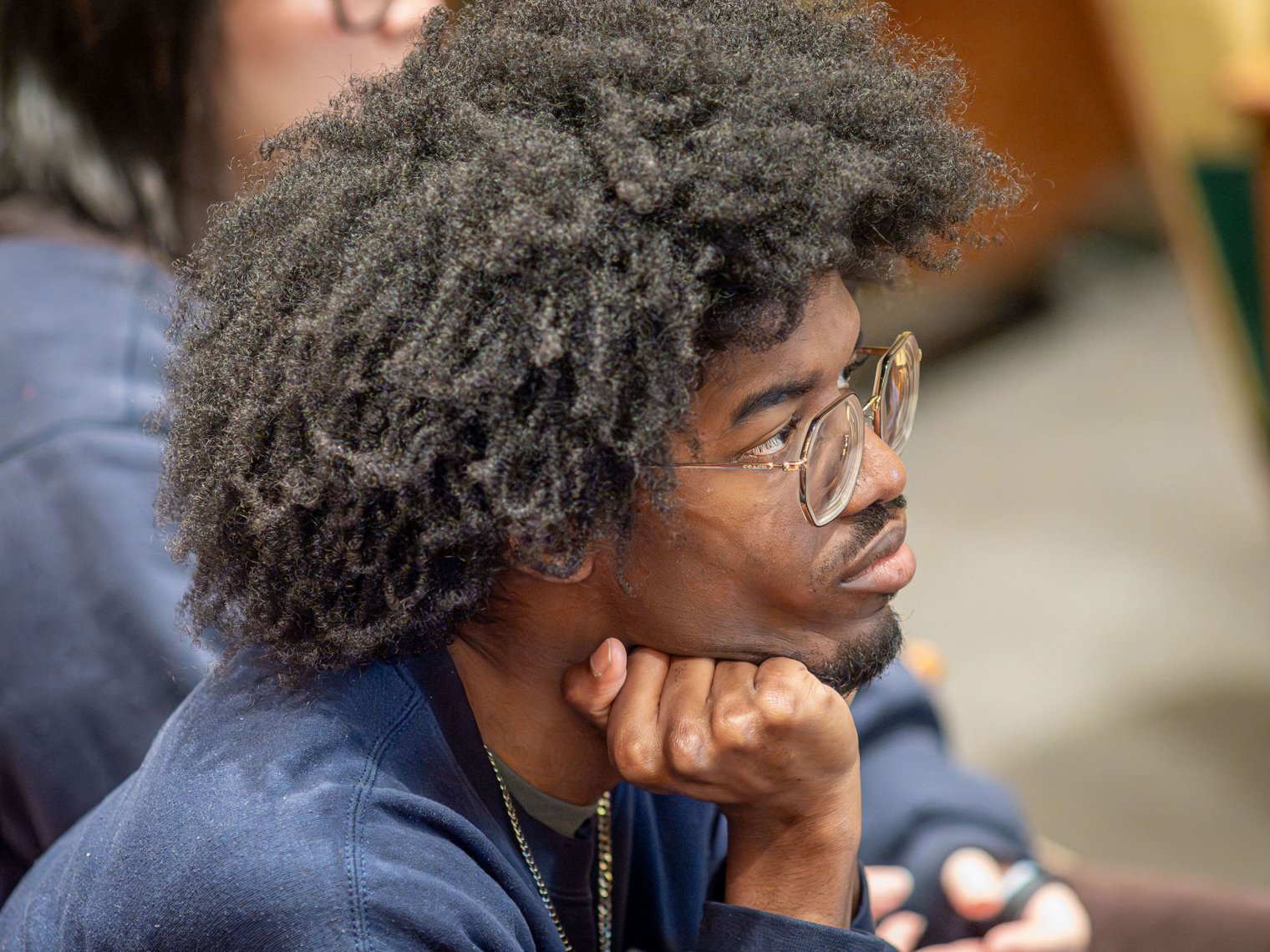 A student wearing glasses holds his hand under his chin in a class.