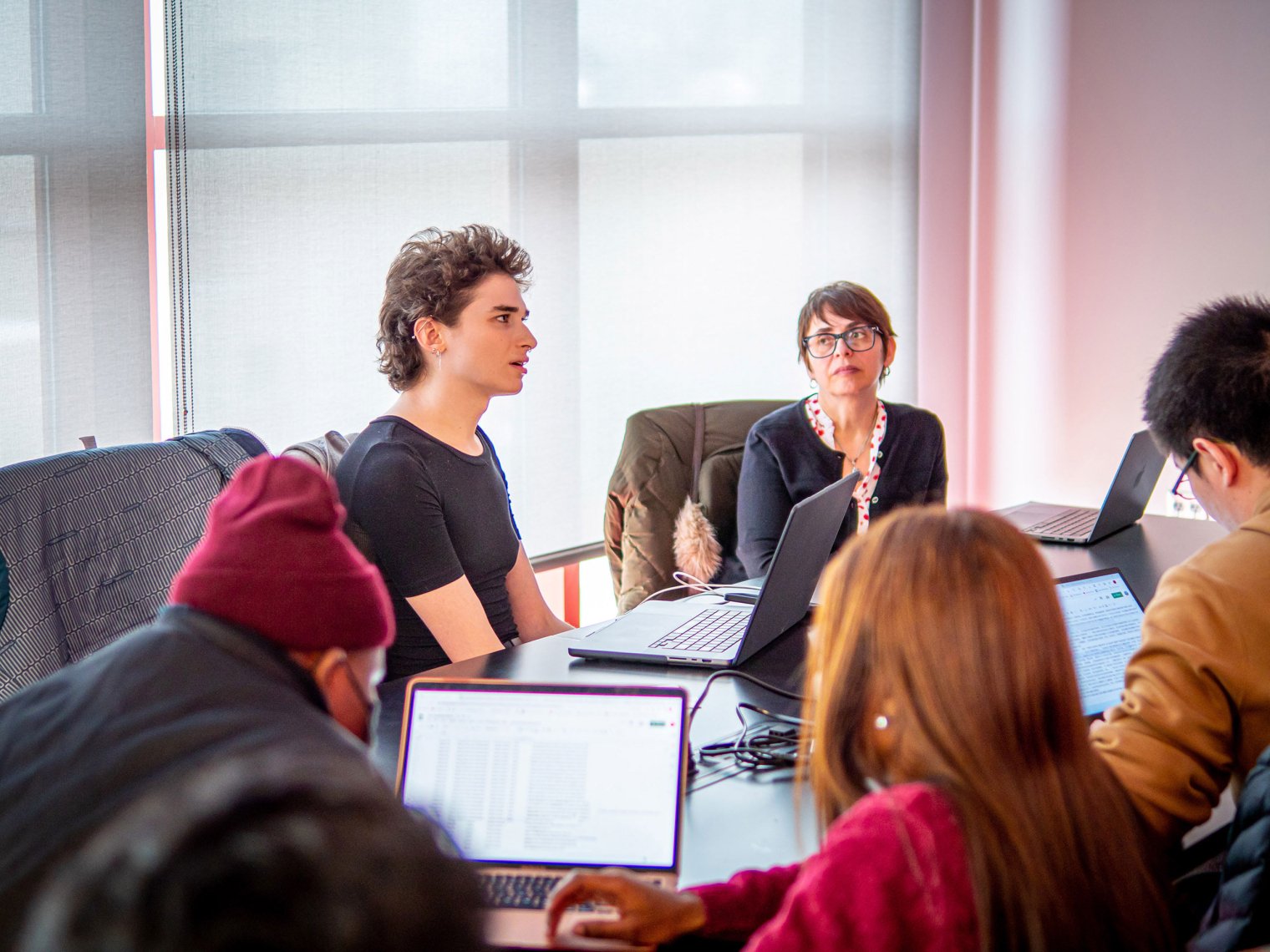 Students sit around a table while a professor looks on in the background.