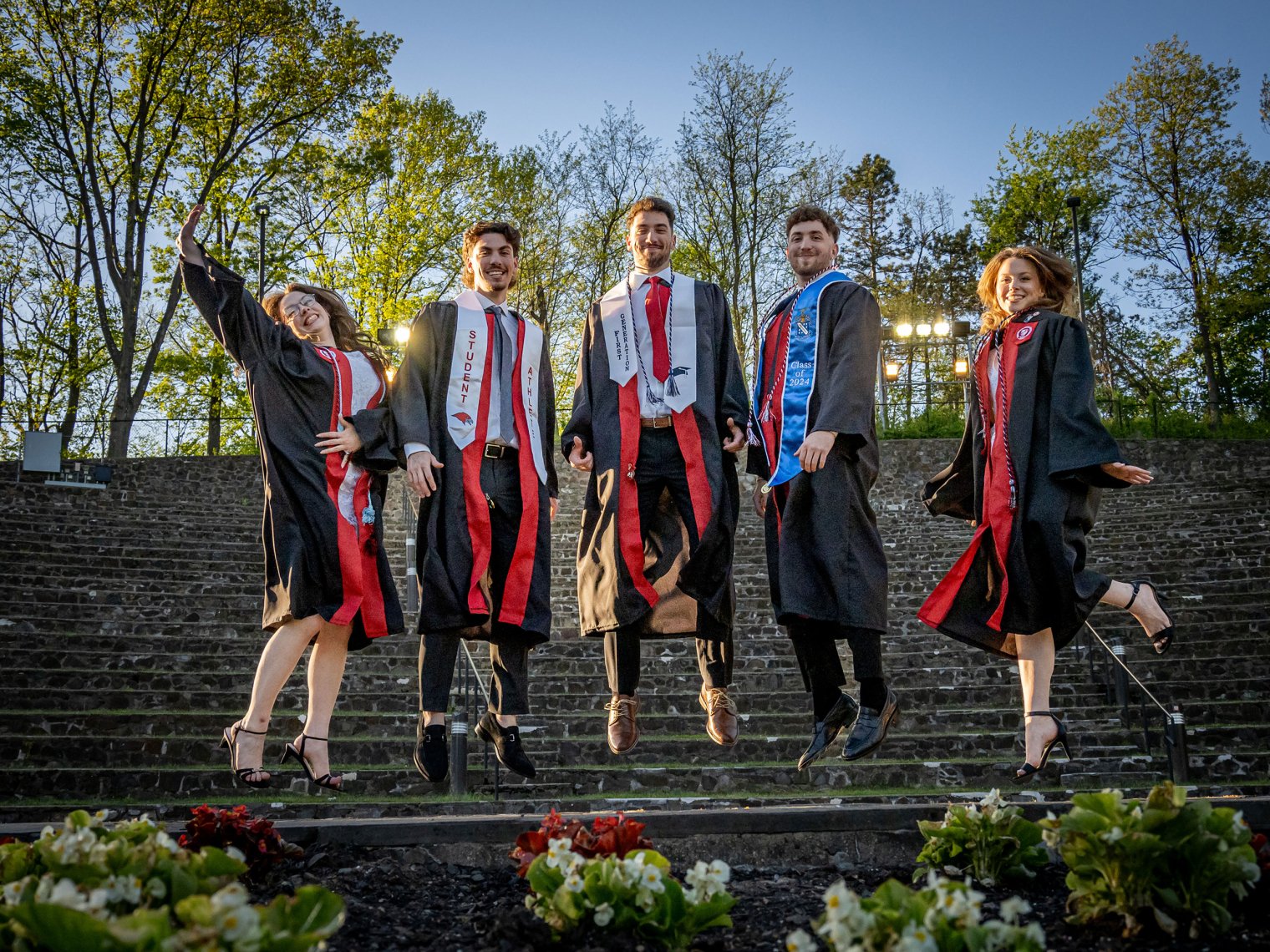 The Povolo quintuplets wearing graduation gowns jump together in front of stone steps outdoors.