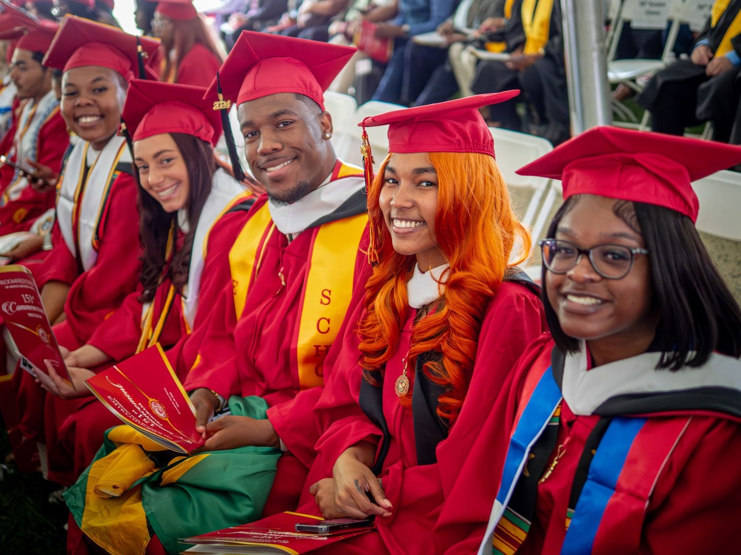 Five students at commencement wearing their cap and gown, sitting down and smiling.