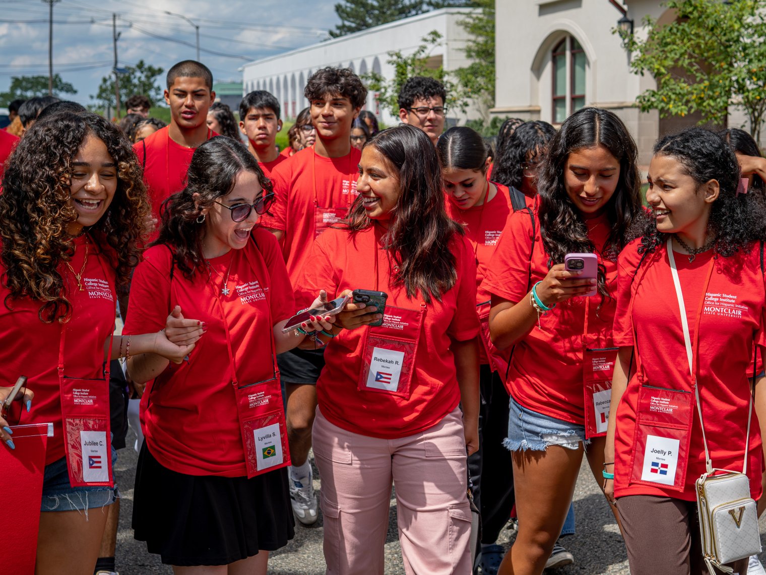A group of students in red t-shirts and lanyards walk together in a large group on college campus.