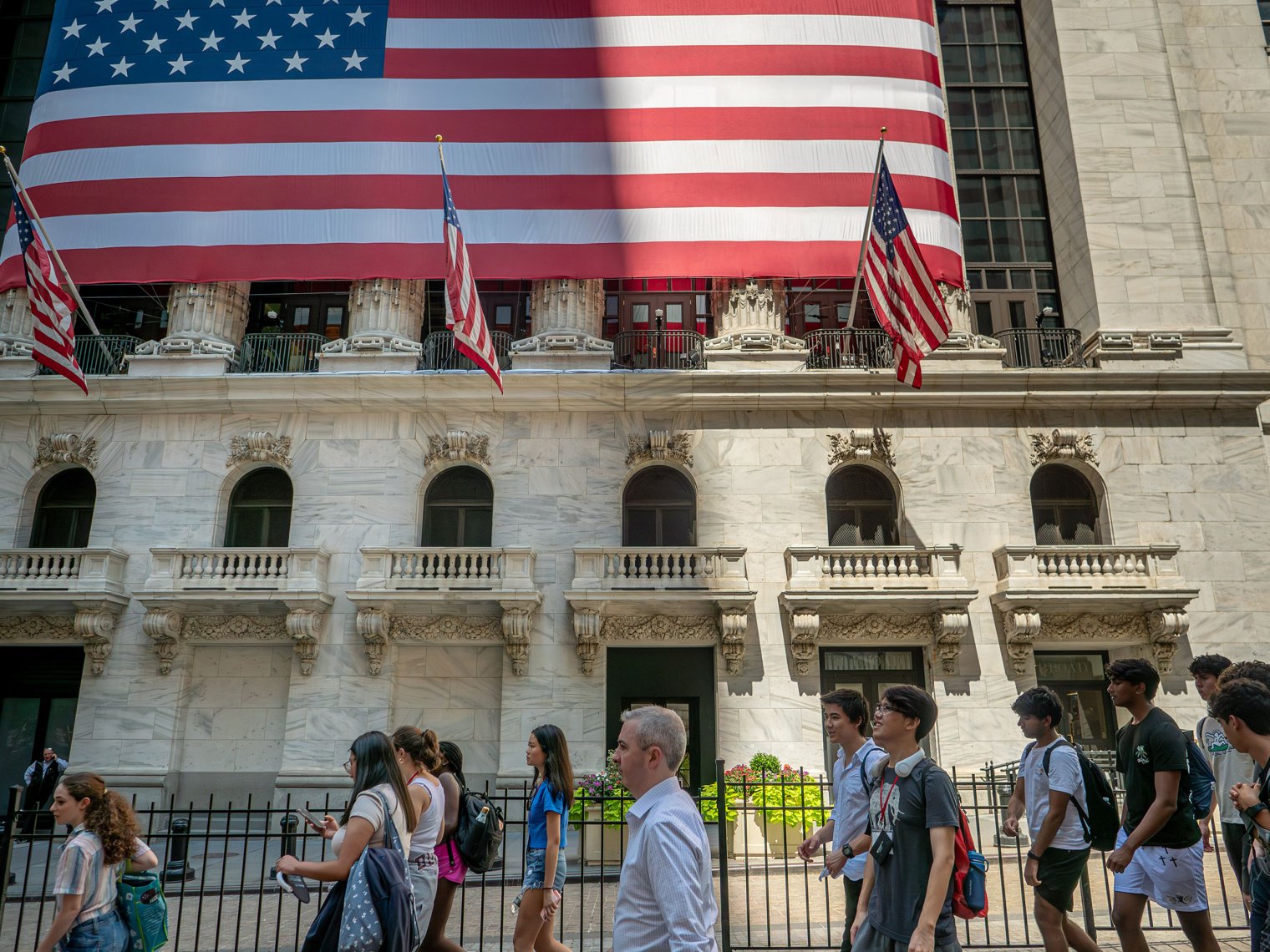 A group of students walks past the New York Stock Exchange in NYC’s Financial District, with a large American flag hanging above the entrance.