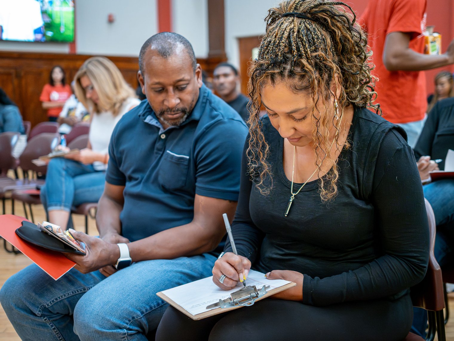 A man and women are next to each other sitting down, the woman is writing down on a clipboard. 