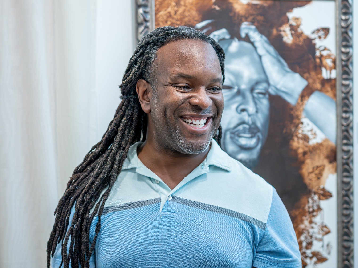 Man is pictured in a blue collared shirt smiling with a painting behind him. 