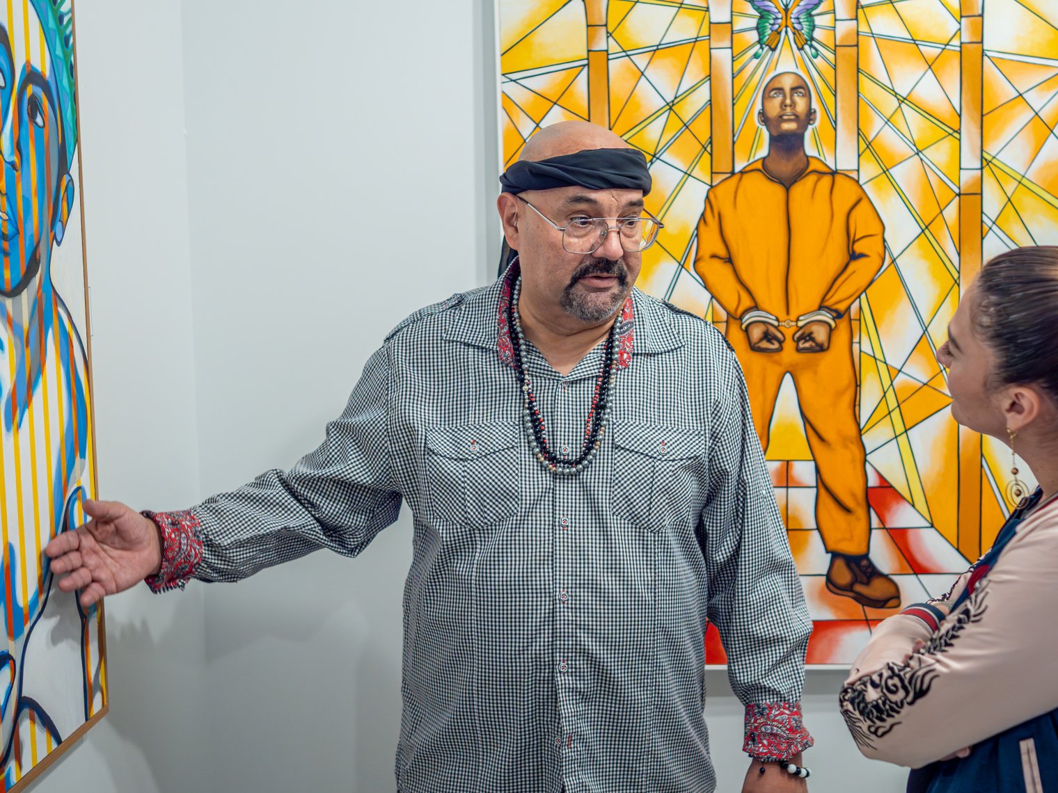 Man is pictured showing a painting to a woman. The main has a black headband, glasses, and a black necklace around his neck. 