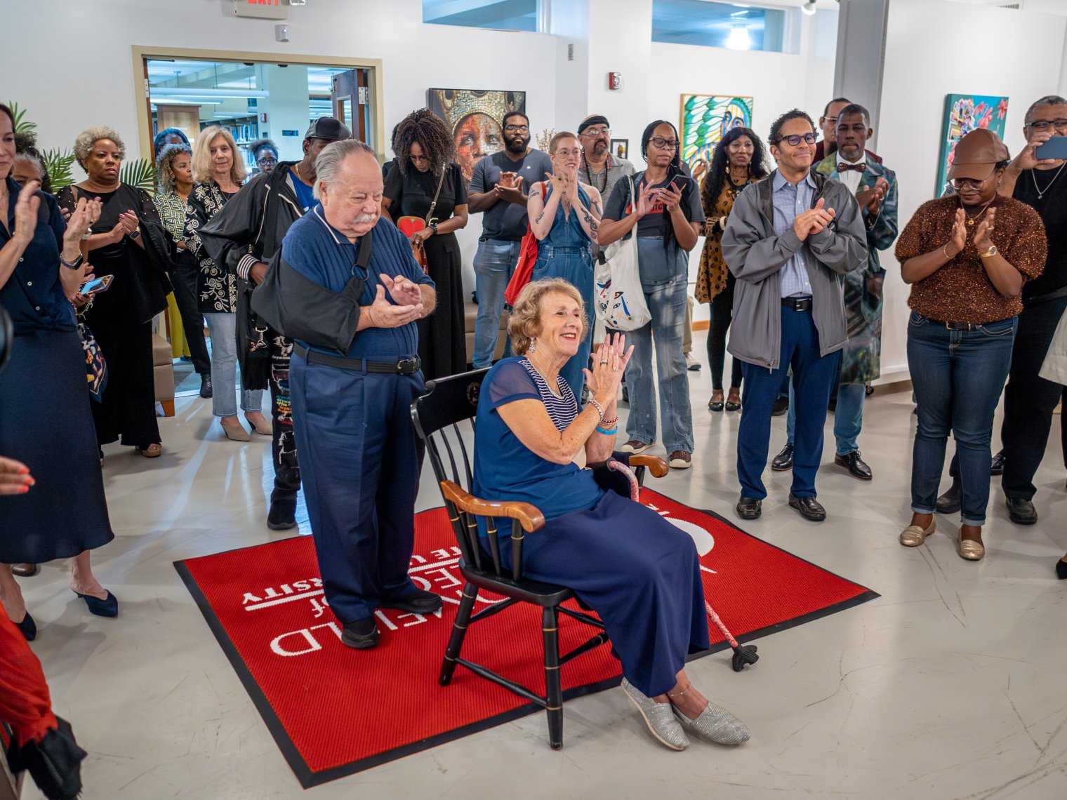 Man is pictured in a cast behind a woman sitting down in a chair, while there is a crowd applauding behind them. 