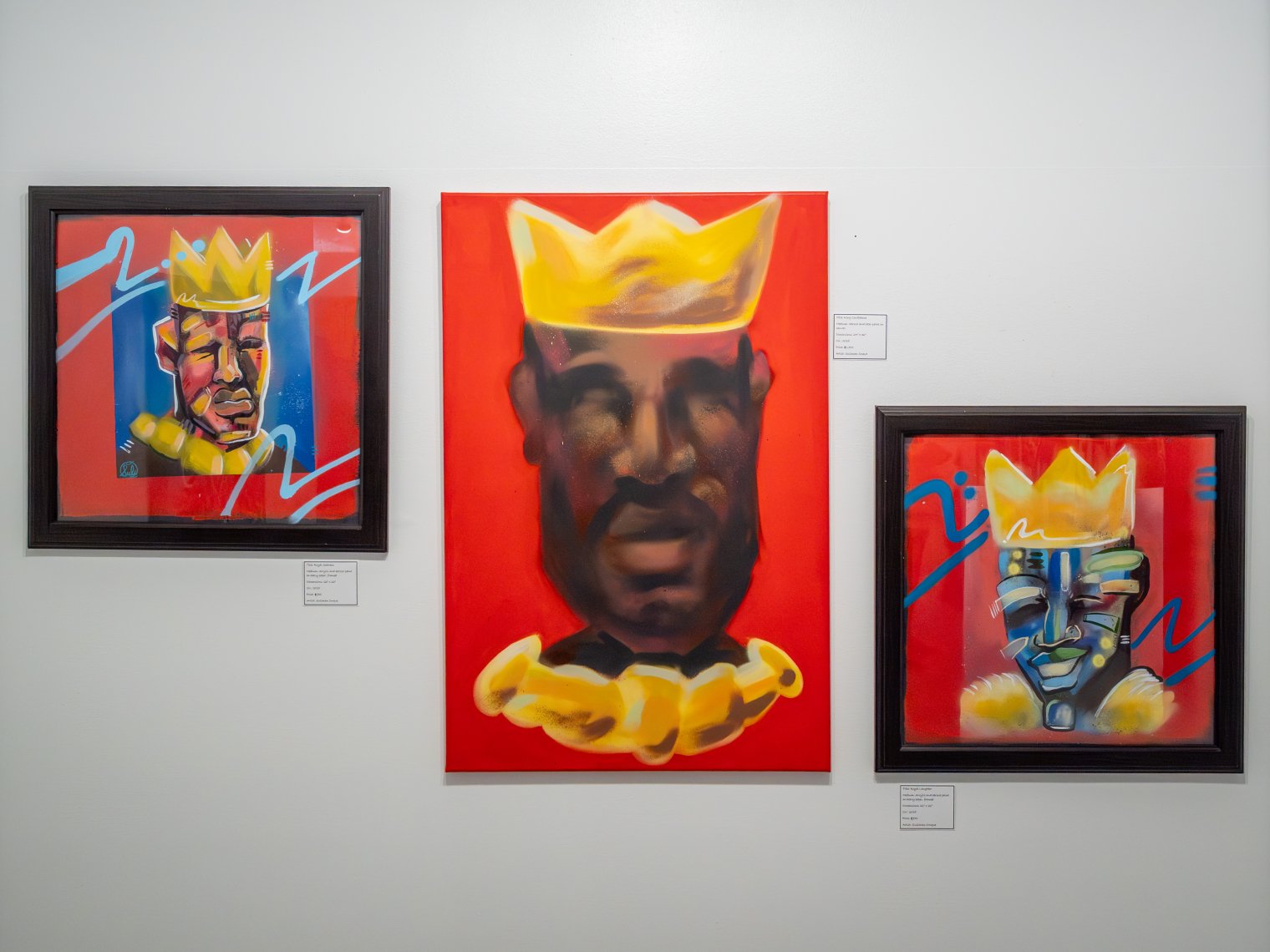 Three paintings are hung on a wall. All three of them with a red background and wearing a gold crown.