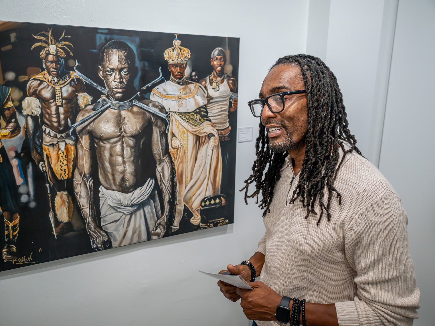 Man is pictured in front a of a painting. Painting consists of five black men in distinct clothing. 
