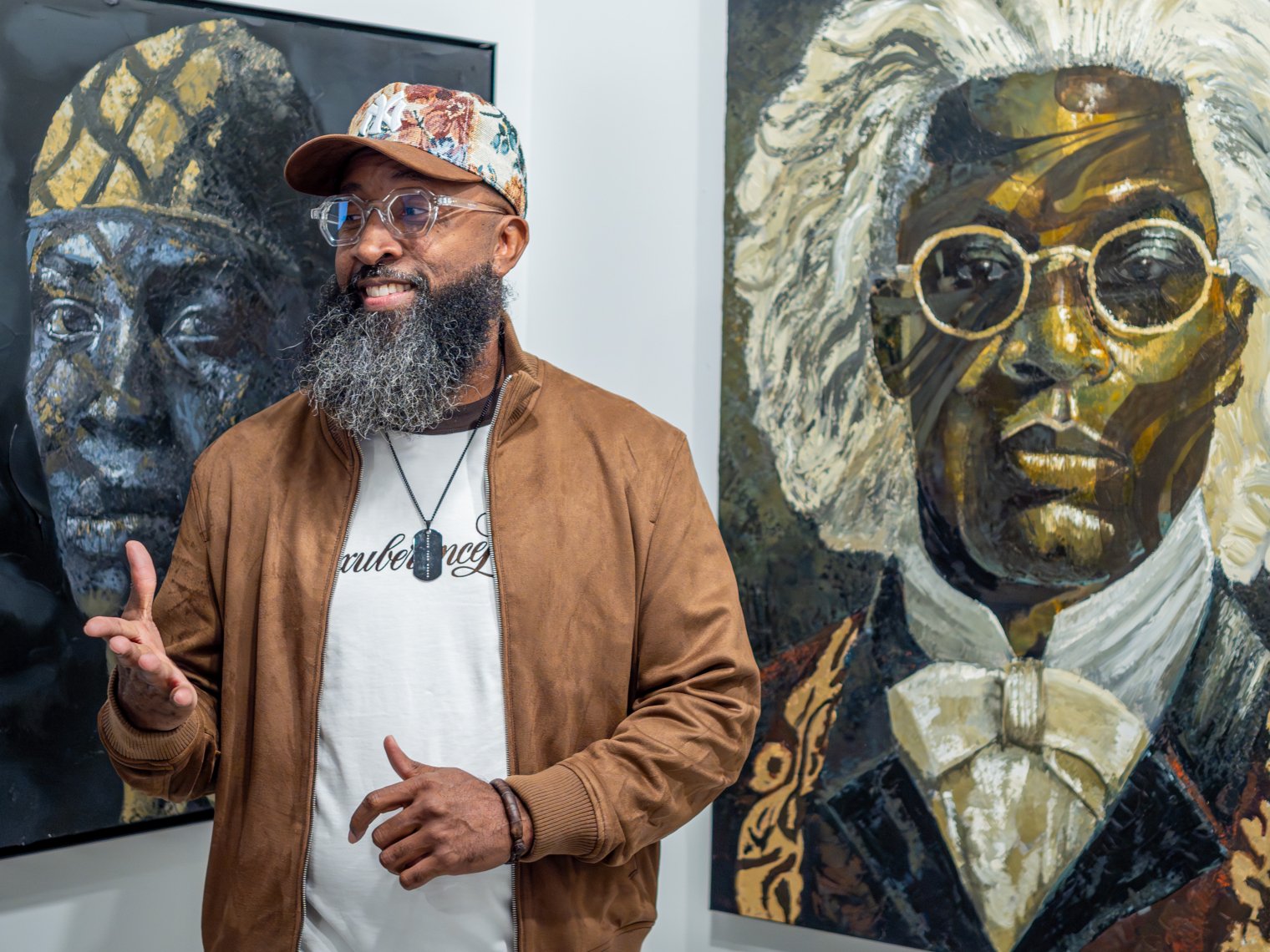 Man is shown in front of two paintings with a hat on and glasses, hand is out as he is explaining something.