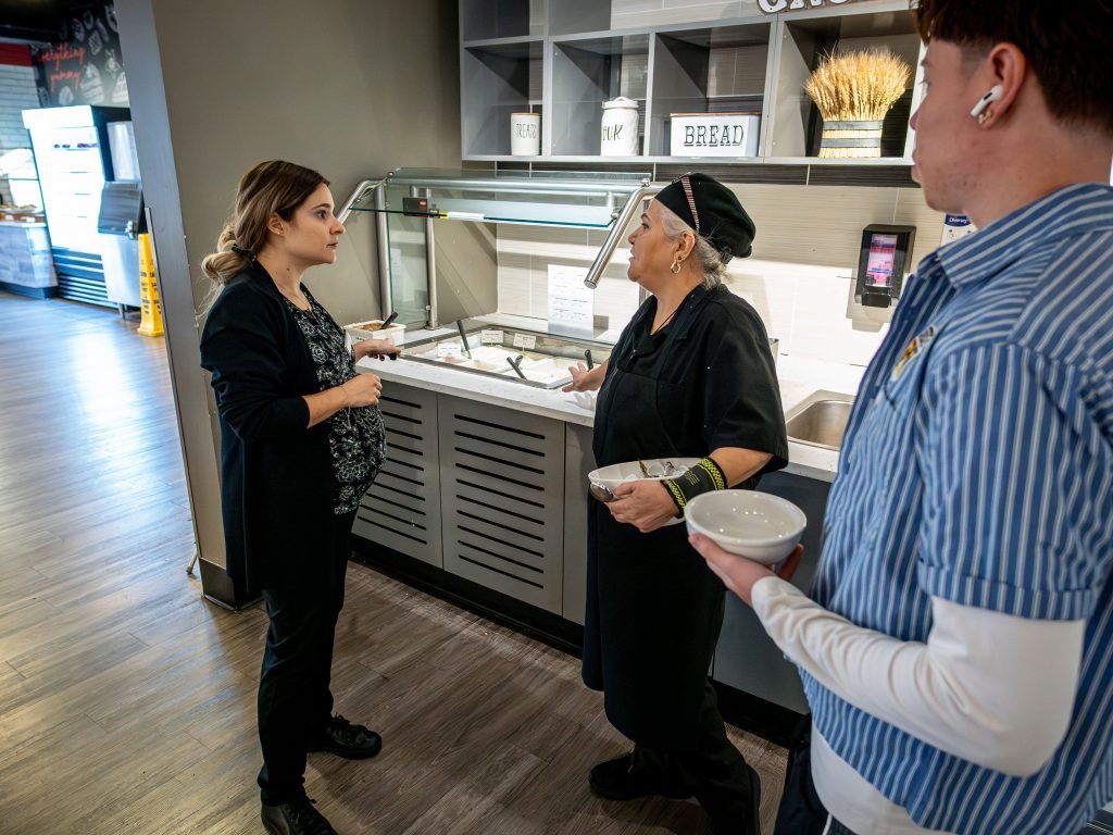 Two woman having a discussion over a hot bar of food, another man is listening intensely. . 