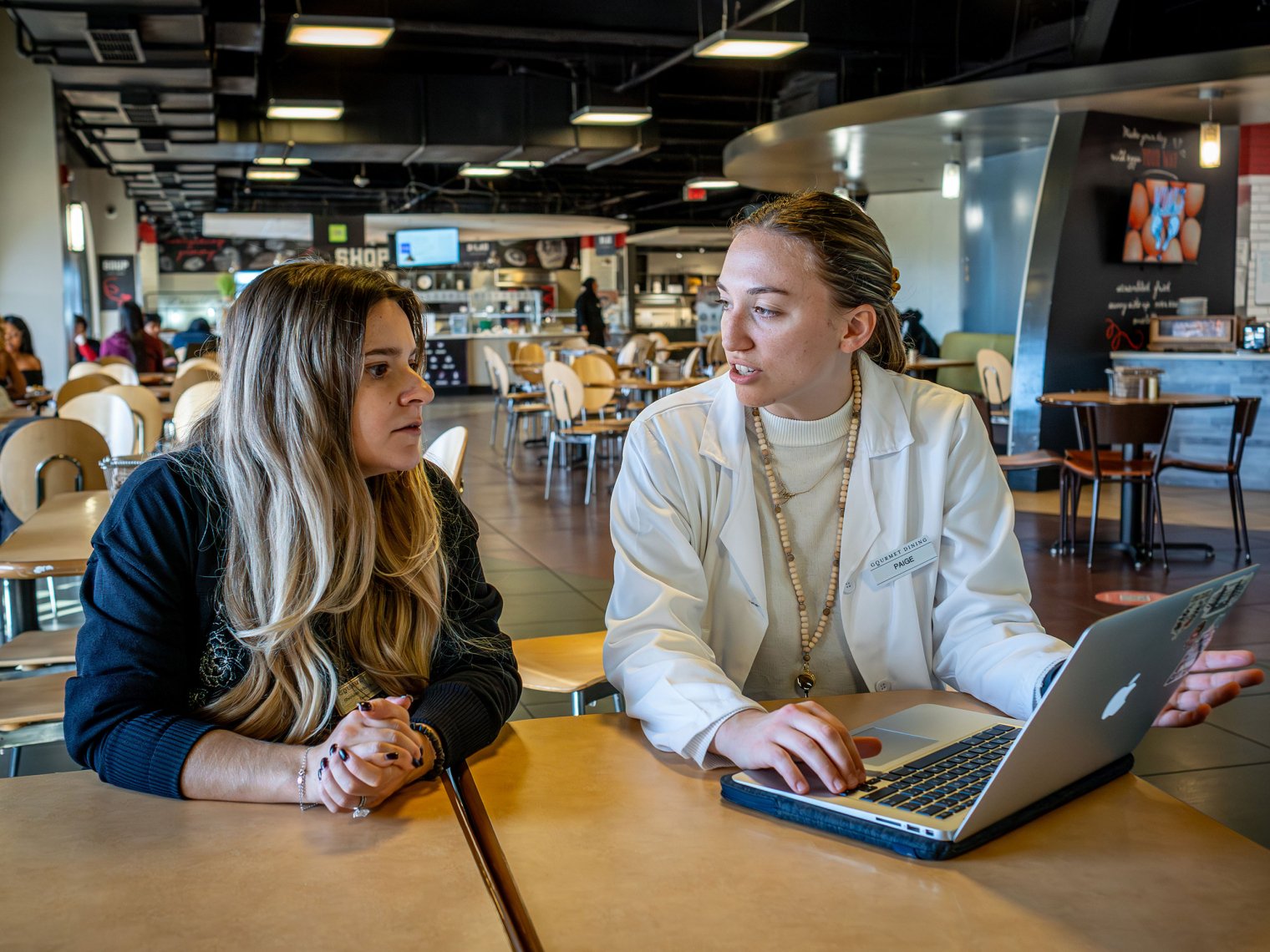 Two women sitting at a desk having a discussion, woman on the right is explaining something on her laptop to the woman on the left. 