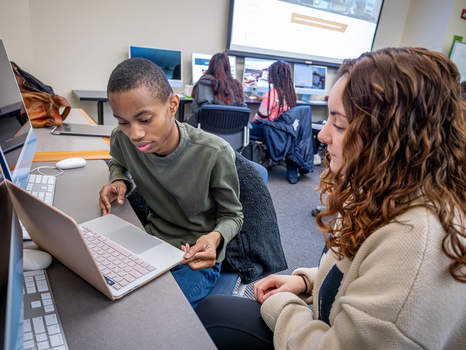 Two students work on a laptop in a classroom, with two more students at a computer behind them.