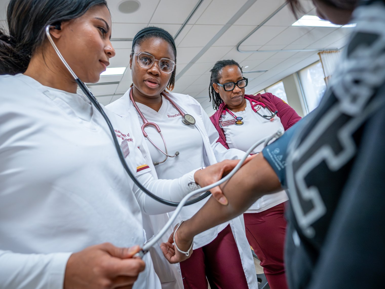 Three woman are pictured in their nursing hear looking over paperwork, one is seen taking the blood pressure of a patient. 