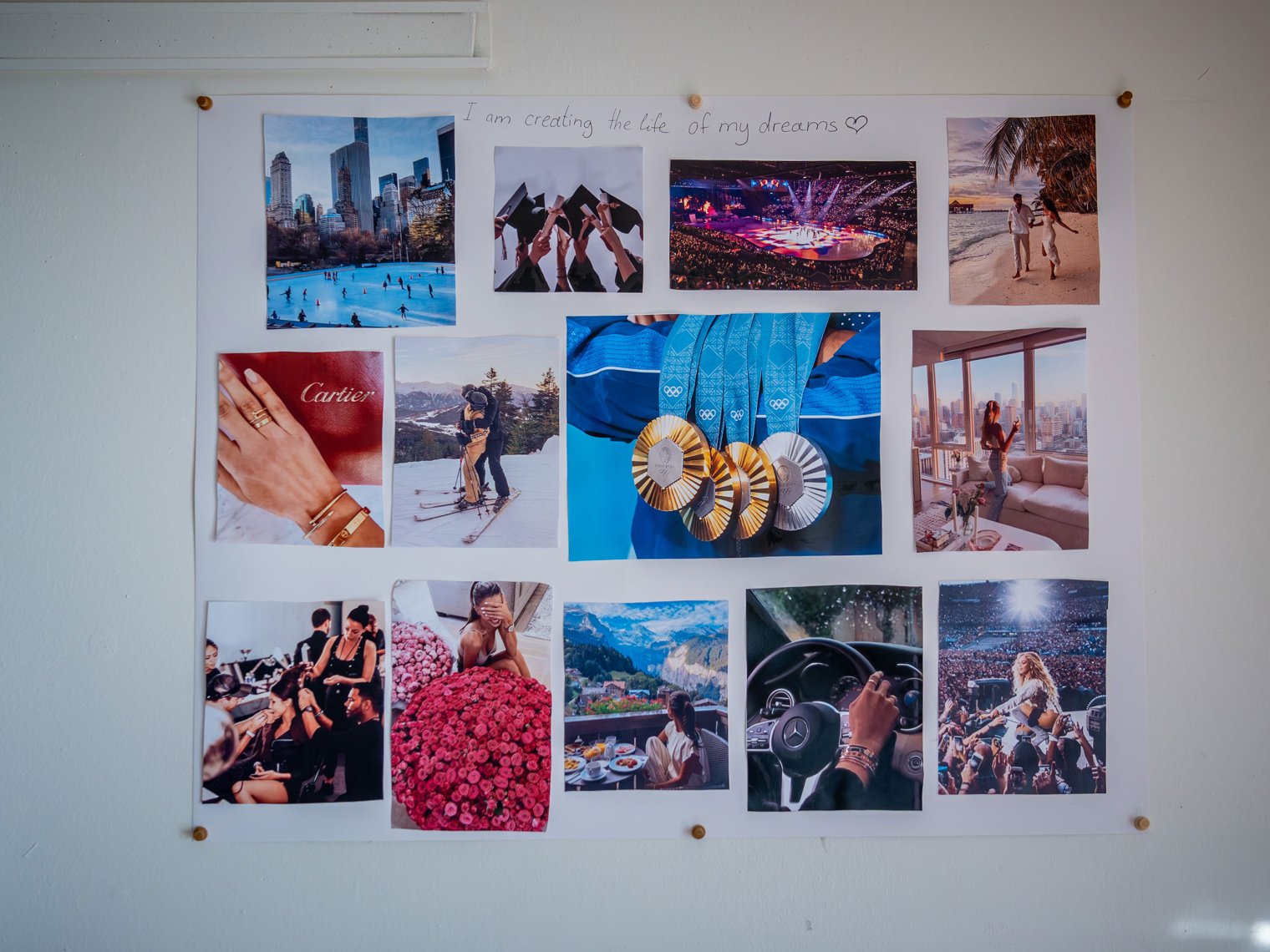 A large vision board hangs on a wall, filled with colorful photos of dream experiences and luxuries, including skaters below a city skyline, graduation caps, Olympic medals, a tropical beach couple. There is a handwritten phrase “I am creating the life of my dreams” across the top.