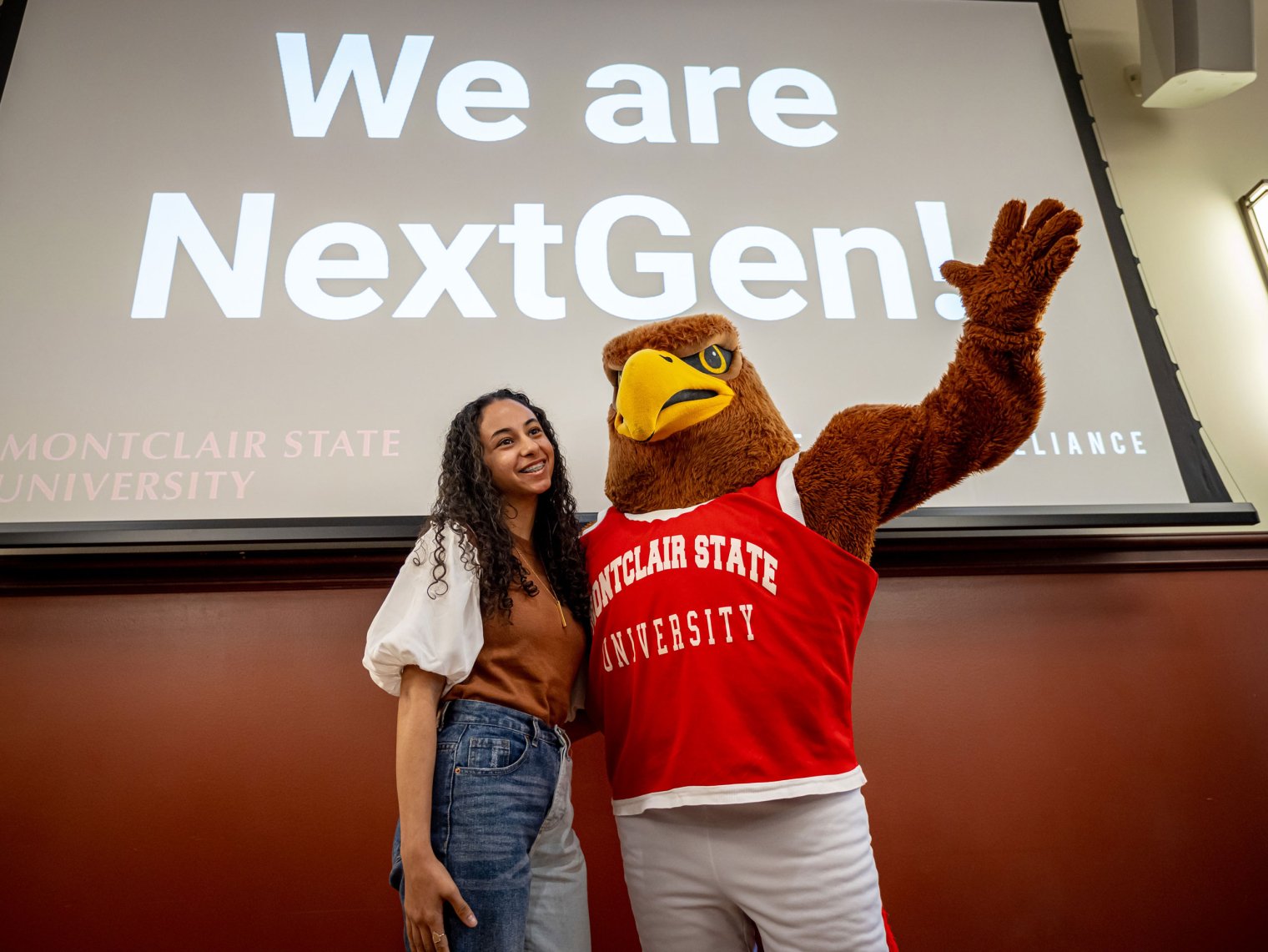 rocky and student standing in front of projector screen displaying a slide that reads We are NextGen