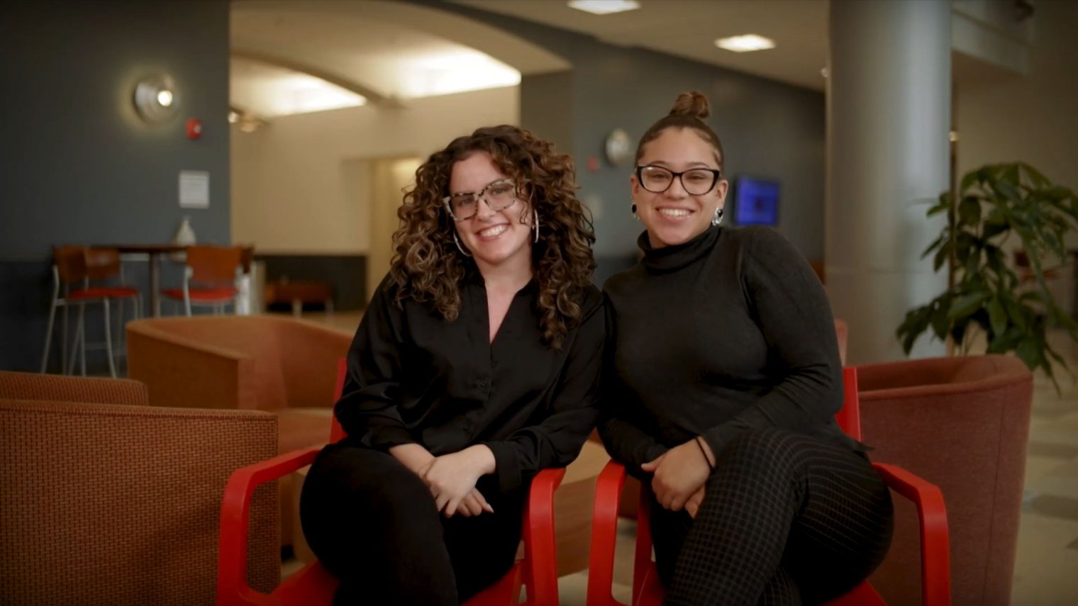 Two women sit in red chairs indoors.