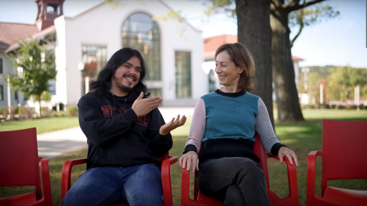 A man and woman sit in red chairs outdoors.