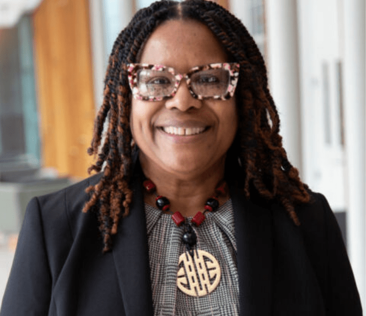 Woman looking directly in camera with leather glasses on, smiling. 