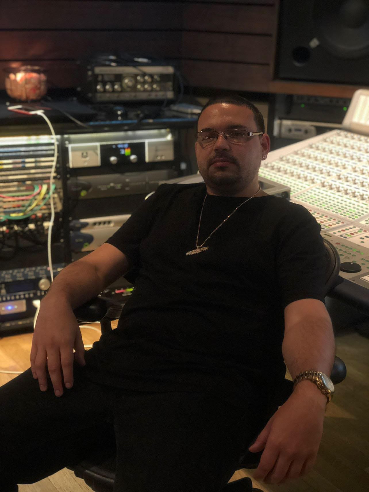 Man is pictured sitting down facing the camera with glasses and a chain on. 