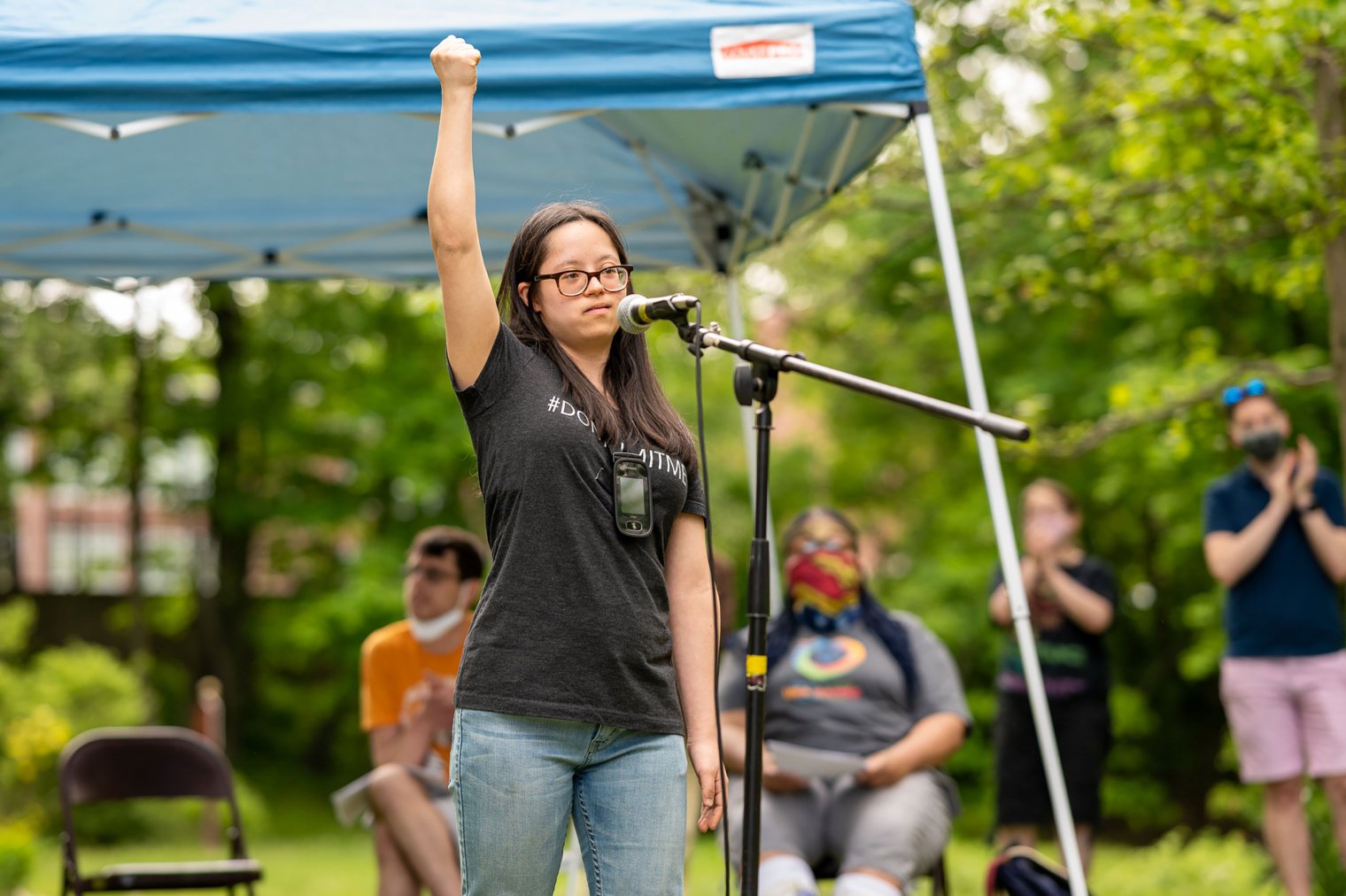 Student raises her fist in the air while standing at a microphone, while people behind her applaud.