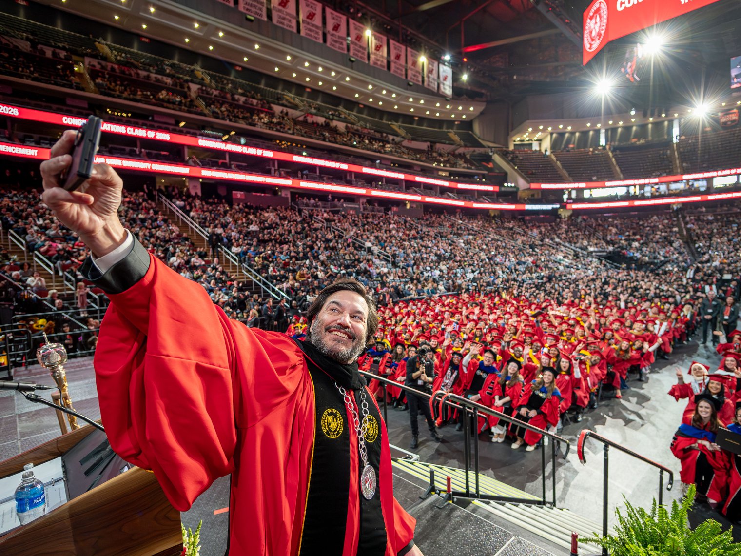Montclair President Jonathan Koppell, wearing academic regalia, stands at the podium and raises his cellphone to take a selfie with a sea of graduates filling the arena behind him.