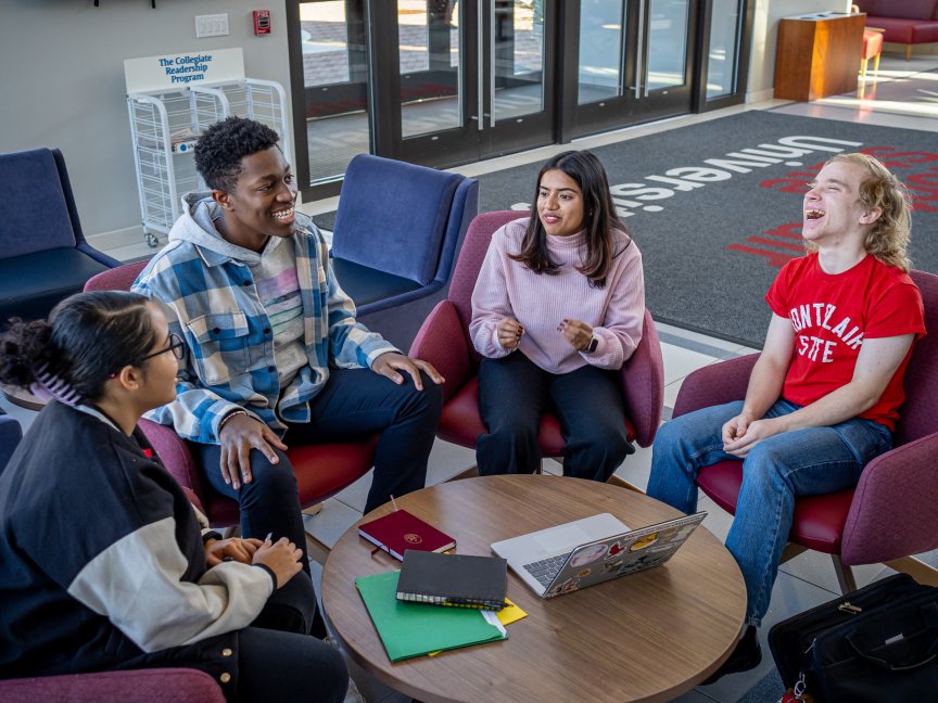 Group of students are sat in front of a round table conversating and laughing with one another.