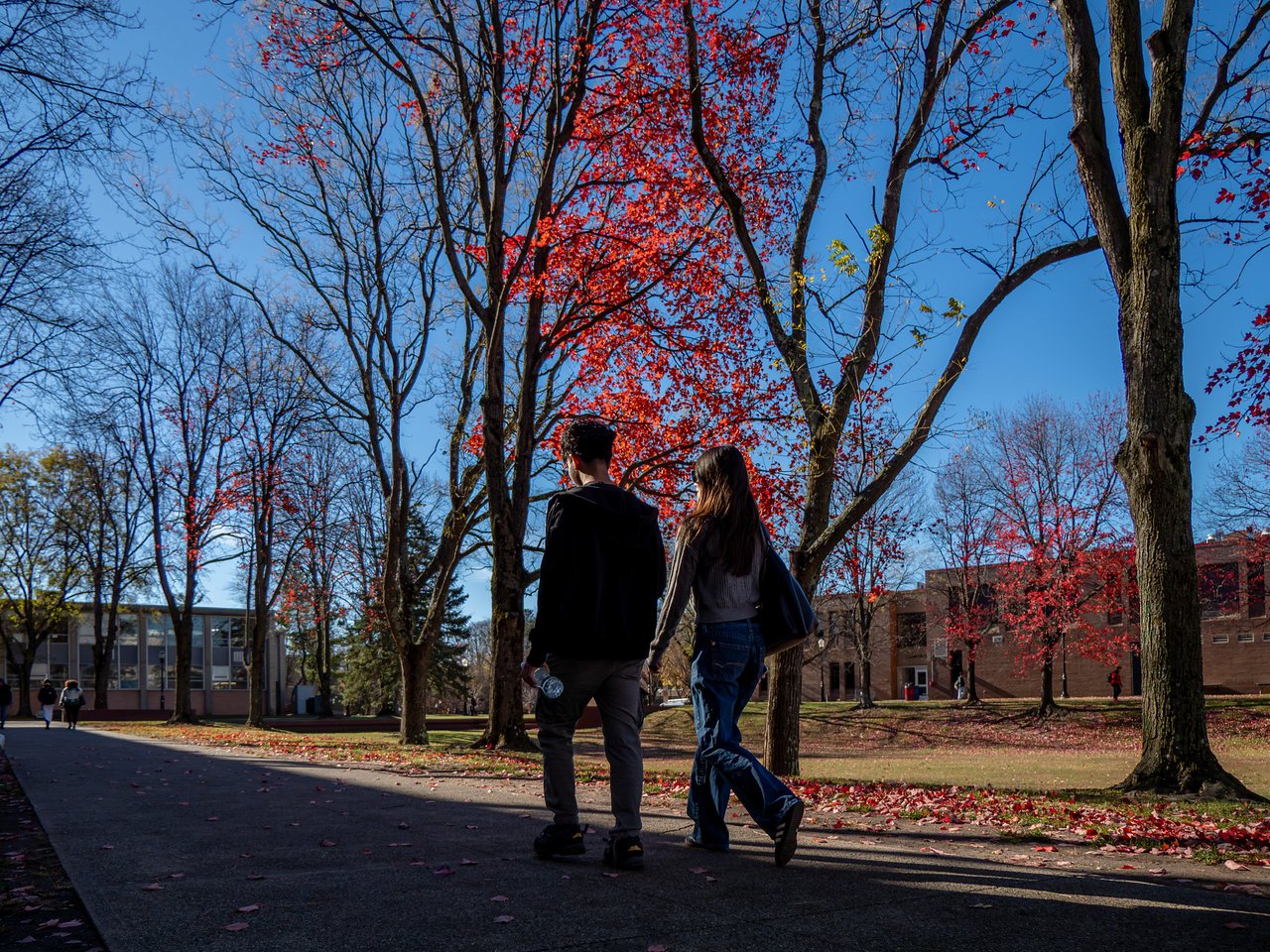 Two students walking through campus with trees with red leaves in the background.