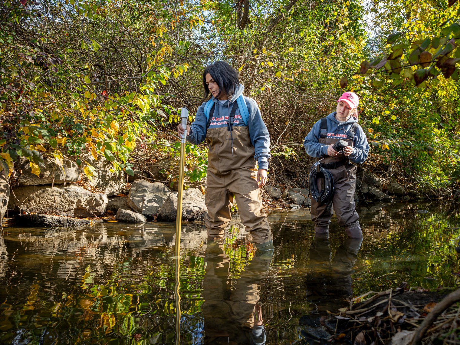 Two Montclair students in waders collecting data in a shallow stream, surrounded by trees and rocks during a field research outing.