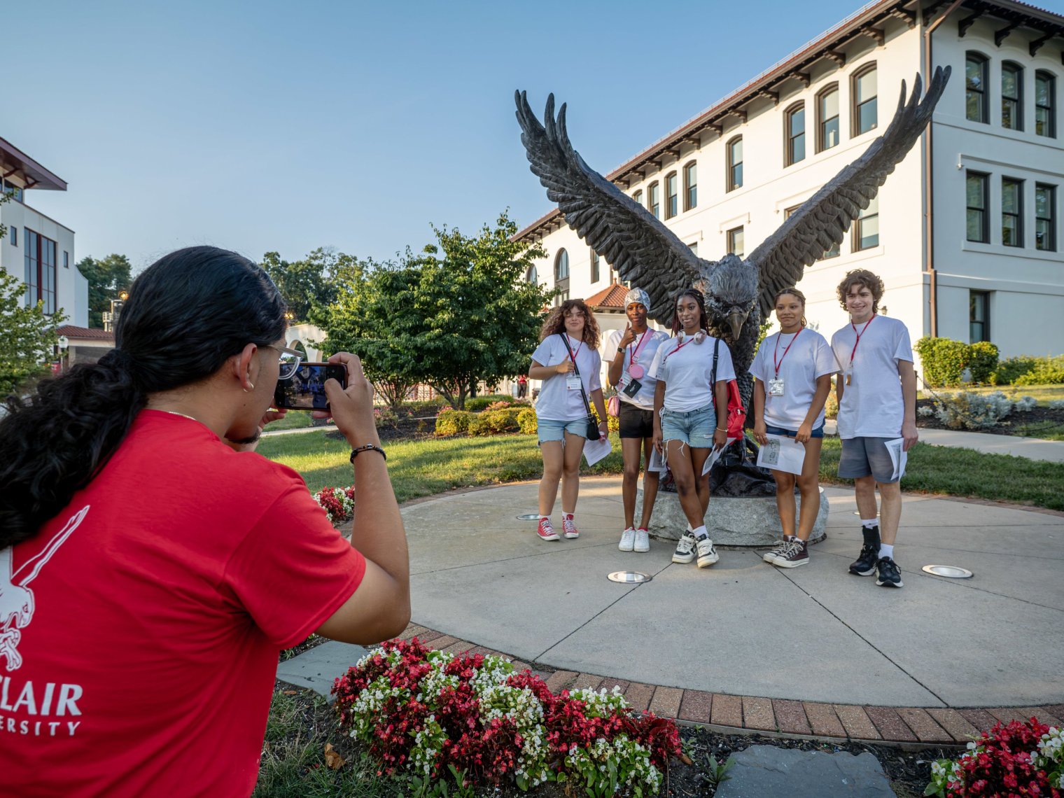 A student ambassador takes a photo of five high school students posing in front of the hawk statue outside Cole Hall