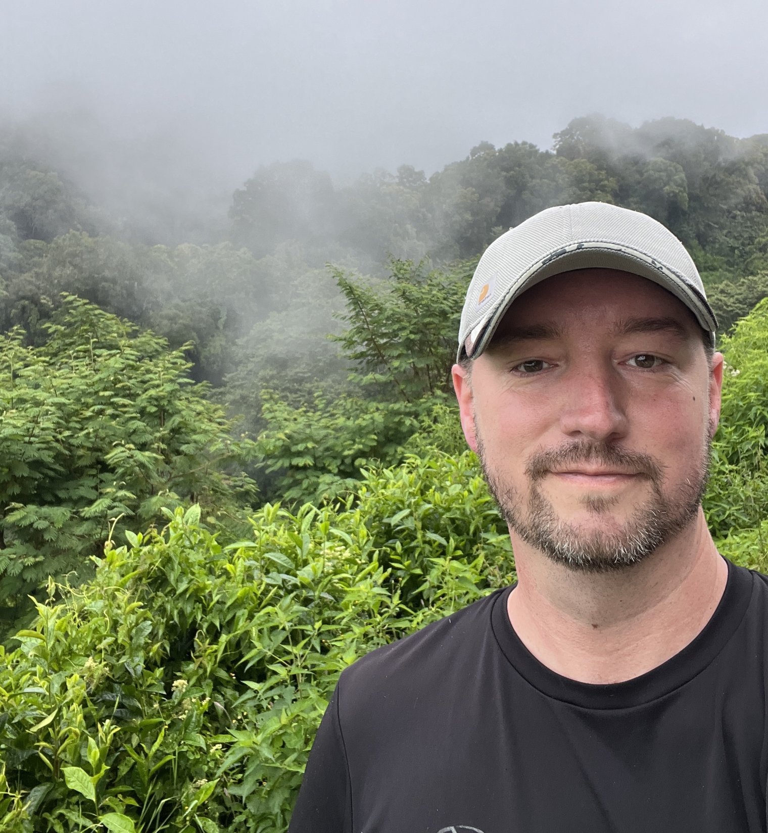 Man smiling with a hat on, behind him green leaves and fog behind him.