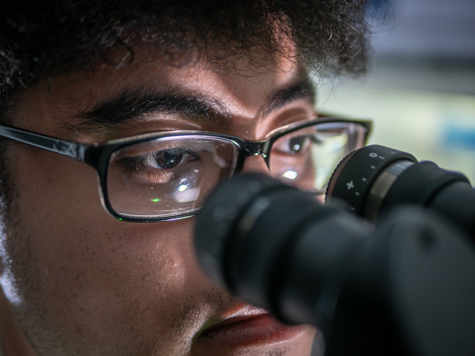 Close-up of Melvin Rodriguez’s eyes behind glasses as he leans into a microscope. The image captures his focused gaze and the reflection of lab light on the lenses.