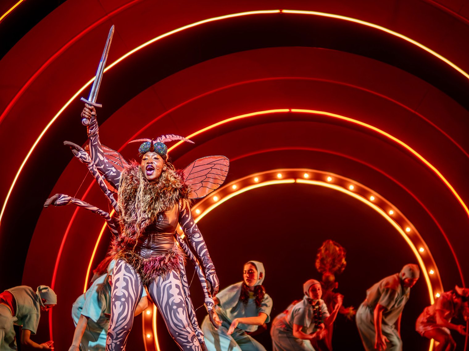 Tenda Kavuna, in character as a giant mosquito, performs and waves a sword onstage as others dance in the background.